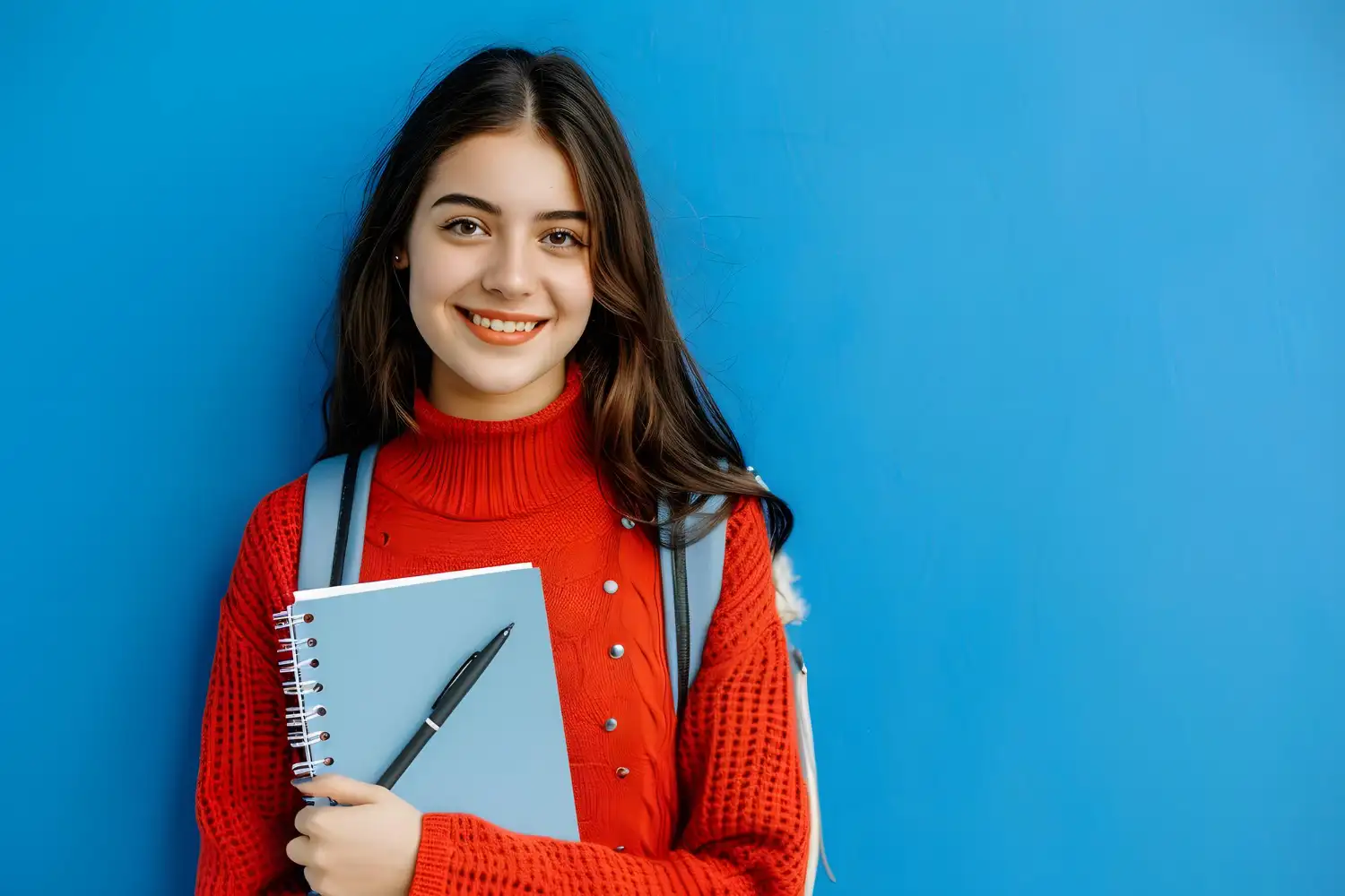 Smiling student in a red sweater holding a notebook and pen, standing against a blue background